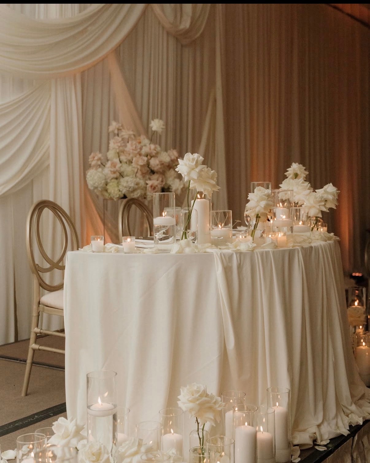 Elegant sweetheart table with a floor-length white linen, white rose and lily floral arrangements, pillar candles, floating candles in glass cylinders, and a draped fabric backdrop at The War Memorial
